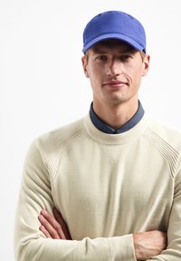 Blauwe stoffen pet met een ronde klep, gedragen met een crèmekleurige gebreide trui met een hoge hals en ribbelaccenten. Model heeft de handen gekruist.