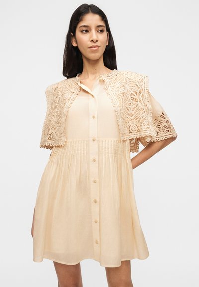 Γυναίκα που φοράει ένα μπεζ φόρεμα με κουμπιά και πιέτες, με έναν μικρό, περίτεχνο δαντελωτό κάππα στους ώμους της, στέκεται μπροστά σε ένα απλό φόντο.