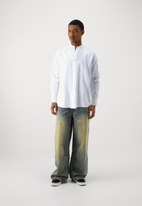 Camicia bianca a maniche lunghe con colletto rialzato, abbinata a jeans in denim blu a gamba larga con una finitura sbiadita e scarpe slip-on nere.