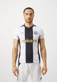 Maglietta da calcio bianca e nera con scollo a V, caratterizzata da un motivo geometrico, testo dorato "nokera" e un logo circolare blu con strisce.