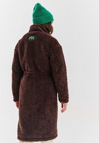 Osoba oblečená v dlouhém hnědém texturovaném kabátu a jasně zelené pletené čepici, stojící zády k fotoaparátu na jednolitém pozadí.