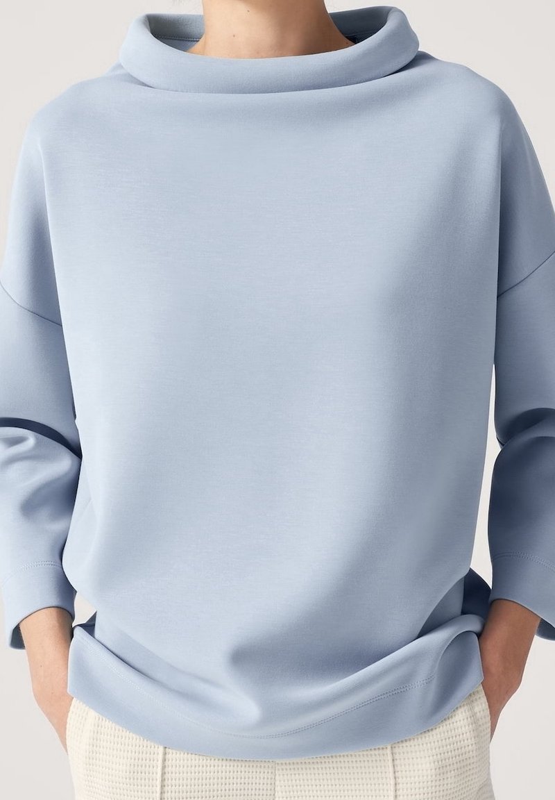 Haut bleu clair à manches longues avec un large col bénitier plié, porté avec un pantalon texturé de couleur claire, mains dans les poches.