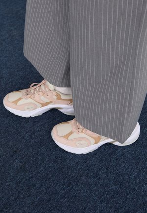 Personne portant un pantalon large gris à fines rayures et des baskets beige-rose, debout sur un tapis bleu foncé.