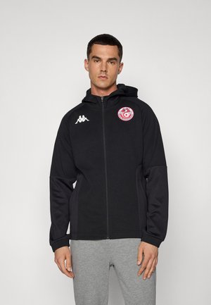 Čierna mikina na zips s kapucňou a kontrastnými rukávmi, logo na hrudníku a štruktúrovaná tkanina. Nosená cez šedé nohavice.