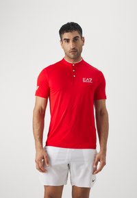 Polo shirt rosso a maniche corte con colletto bottonato e dettagli bianchi, caratterizzato da un logo EA7 sul petto e un design sportivo.