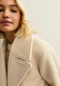 Mujer rubia vistiendo un abrigo de lana color crema con grandes solapas y un collar de oro, contra un fondo claro.