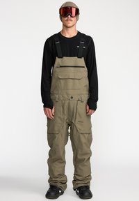 Khaki waterdichte bretelsneeuwbroeken met verstelbare banden, meerdere zakken en versterkte manchetten. Zwart langärmelige shirt en een bril gedragen.