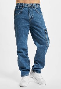 Blaue Denim-Cargo-Hosen mit einer lockeren Passform, ausgestattet mit Seitentaschen, einem Knopfverschluss und weißen Sneakers. Der Stoff hat eine strukturierte Oberfläche.