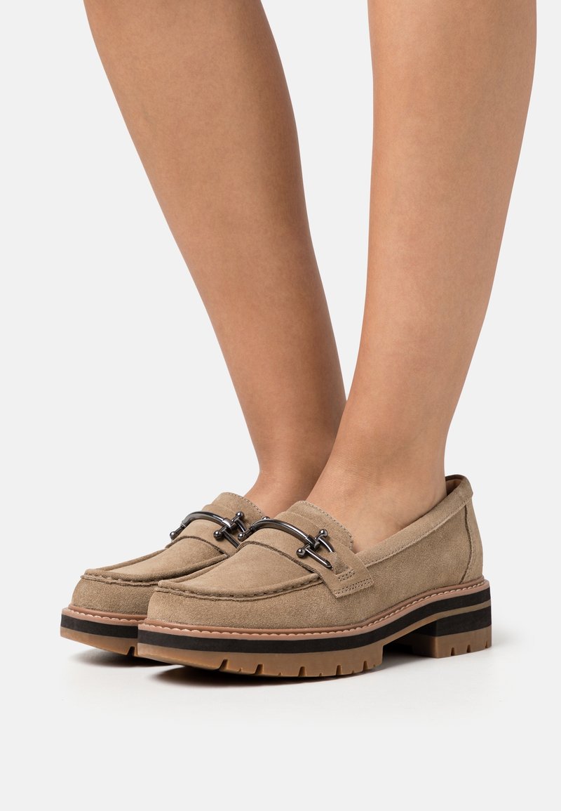 Des mocassins en daim beige présentent un bout arrondi, des accents de quincaillerie noire et une semelle extérieure en cuir brun épais avec des motifs de traction robustes.