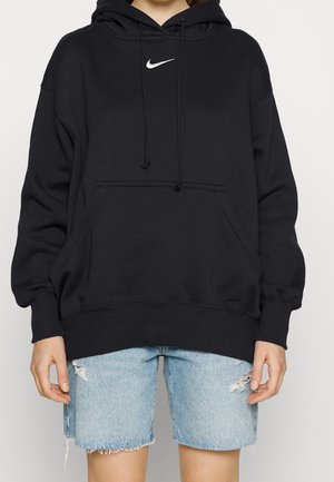 Osoba ubrana w czarną bluzę Nike z kapturem i kieszenią z przodu oraz jasnoniebieskie, podarte szorty jeansowe na jednolitym tle.