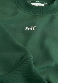 Zielona bluza z miękkiego materiału, z prążkowanym kołnierzem i mankietami. Na przodzie haftowane białe logo "self.".
