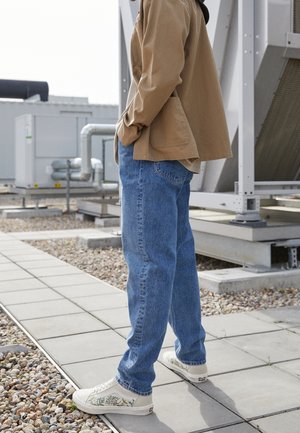 Osoba w niebieskich dżinsach, beżowej kurtce i białych trampkach z kwiatowym wzorem stoi na wyłożonym kafelkami dachu w pobliżu sprzętu przemysłowego.