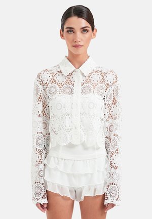 Frau trägt eine weiße Spitzenbluse mit langen Ärmeln und Blumenmuster sowie weiße, geraffte Shorts und steht vor einem einfarbigen Hintergrund.