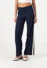 Un pantalon large bleu marine avec une bande latérale beige, fabriqué en tissu léger, avec une texture lisse et une coupe décontractée.