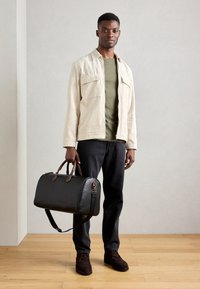 Zwarte leren duffelbag met bruine leren accenten, zachte textuur, cilindervorm en een afneembare schouderband; model draagt een beige cordjack.