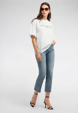 Mujer con camiseta blanca con gráfico, jeans acampanados azul claro, sandalias negras de tacón alto y gafas de sol marrones, de pie con una mano en el bolsillo.