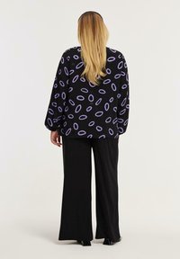 En blond kvinde vender væk, iført en sort bluse med lilla oval mønstre, parret med bredbenede sorte bukser og sorte hæle.