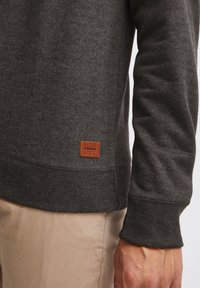 Donkergrijze sweatshirt van zacht materiaal, met ribbels aan de mouwen en een gestructureerde tailleband. Een bruine leren label met "BLEND" is zichtbaar.