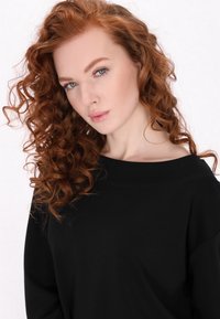 Top nero, ampio e con design a spalle scoperte. La modella ha lunghi capelli ricci rossi e trucco delicato, con uno sfondo chiaro e uniforme.
