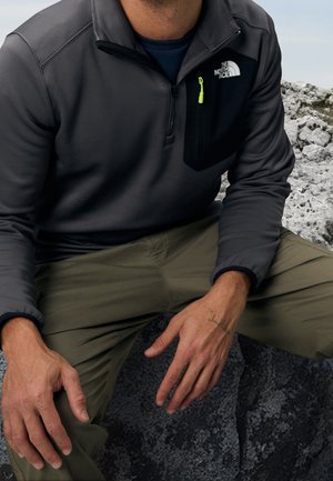 Maglione grigio con zip a quarto realizzato in tessuto morbido, con una tasca sul petto nera e una zip gialla neon. Pantaloni oliva e sfondo roccioso.