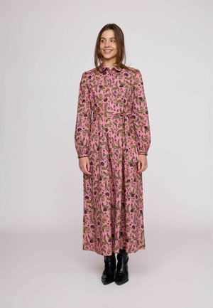 Giovane donna in piedi che sorride, indossa un lungo vestito rosa a fiori e stivaletti neri su uno sfondo chiaro e uniforme.