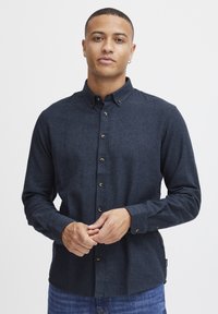 Camisa de botón azul oscuro hecha de tela texturizada, con cuello, detalles de botones y mangas largas. Se lleva con jeans de mezclilla.