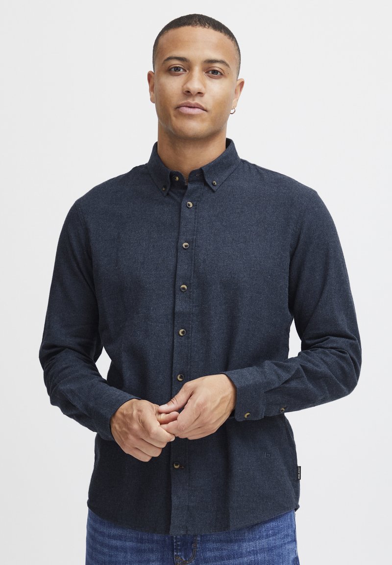 Camisa de botón azul oscuro hecha de tela texturizada, con cuello, detalles de botones y mangas largas. Se lleva con jeans de mezclilla.