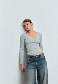 Svjetlosivi rebrasti top dugih rukava sa valovitim izrezom, upareni s visokim strukom plavim trapericama. Glatka tekstura i uski kroj.