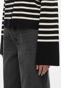 Persona che indossa un maglione a maniche lunghe a righe nere e crema con polsini larghi e jeans di denim neri sbiaditi.