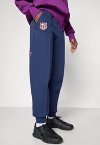 Pantalons de survêtement navy avec un logo de Barcelone, poches latérales et poignets élastiques, associés à des baskets noires avec un accent vert.