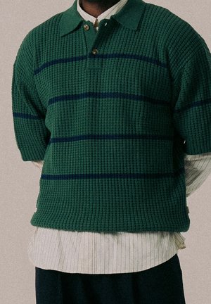 Persona vistiendo un suéter de punto verde de manga corta con rayas oscuras sobre una camisa de cuello a rayas finas claras y pantalones oscuros.