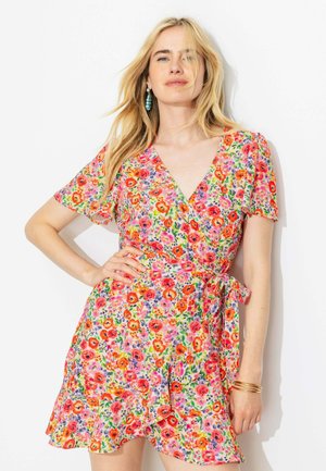 Femme blonde portant une robe portefeuille courte à fleurs rouges, roses et violettes, des boucles d'oreilles turquoise et des bracelets en or, posant devant un fond blanc.