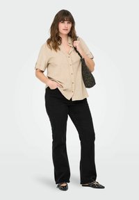 Camisa de botones de manga corta beige, jeans de campana negros y zapatos planos negros con acentos de tachuelas. La modelo lleva un bolso de estampado de leopardo negro y marrón.