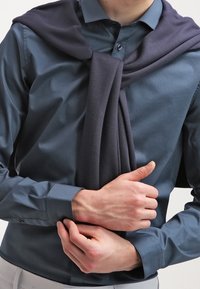 Tmavě modrá košile s dlouhými rukávy, manžetami na knoflíky, a pletený námořnický svetr přehozený přes ramena, s měkkou texturou a záhyby.