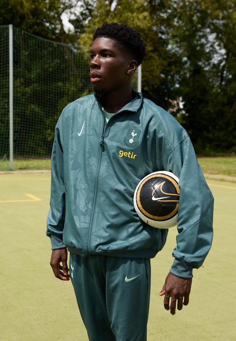 Giacca sportiva verde e pantaloni con marchio Nike; tiene in mano un pallone da calcio nero e oro. Sullo sfondo si vede un campo di calcio con una porta.