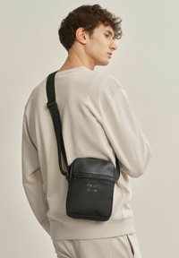 Czarna torba na ramię z gładkiego materiału, z regulowanym paskiem, kieszenią na zamek i wytłoczonym tekstem. Kompaktowy prostokątny kształt.