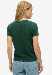 Blonde vrouw die van zich af kijkt, gekleed in een donkergroen T-shirt met korte mouwen en lichte spijkerbroek, tegen een eenvoudige witte achtergrond.