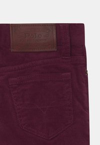 Burgundské kordové kalhoty s koženou nášivkou „Polo Ralph Lauren“. Jsou viditelné detaily kapes a vertikální vroubkovaná textura.
