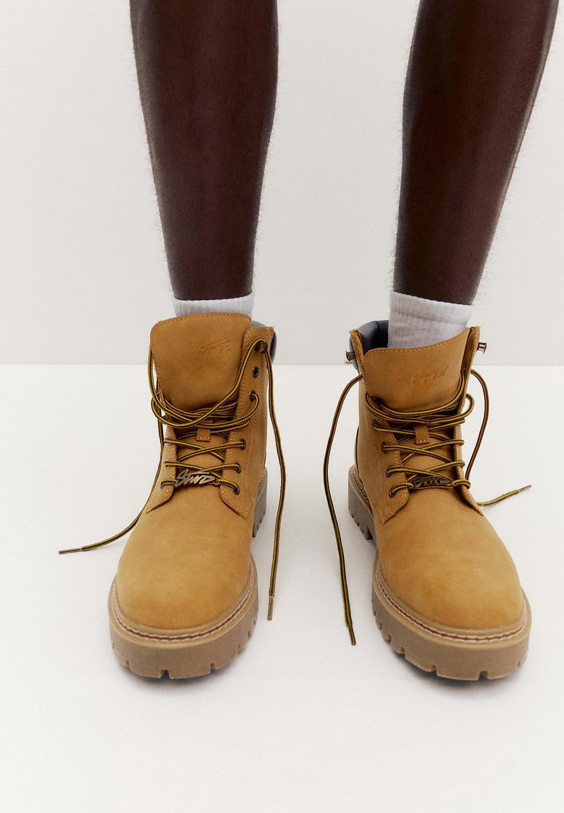 Osoba koja nosi bijele čarape i smeđe gležnjače s debelim potplatima i vezicama, stoji na običnoj bijeloj površini na neutralnoj pozadini.