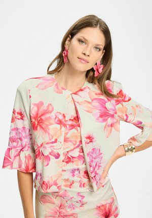 Kvinde med lyst brunt hår iført en lys beige blomsterjakke og kjole med pink blomster, matchende pink blomst øreringe og en guldbelagt armring.