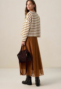 Femme portant une jupe en dentelle marron, un haut à rayures blanches et marron à manches longues, des bottes noires et tenant un sac à main marron foncé avec des œillets en métal.