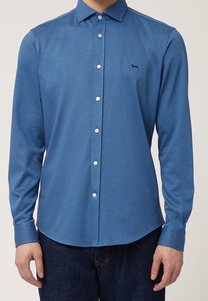 Mężczyzna ubrany w niebieską koszulę na guziki z długim rękawem, z małym ciemnym haftowanym logo psa na piersi.