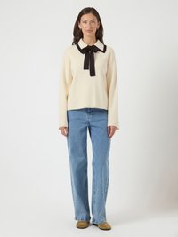 Mulher de pé, vestindo um suéter creme com gola e laço pretos, jeans azul claro e sapatos bege, contra um fundo branco.