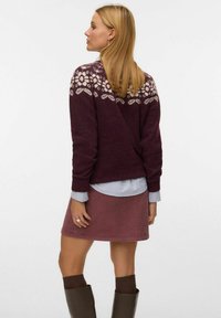 Un pull en laine bordeaux avec un motif tricoté floral sur les épaules, associé à une jupe en velours côtelé rose. Des bottes brunes montantes et une chemise superposée.