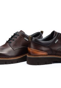 Chaussures en cuir marron avec bout rond, surface texturée, accents marron contrastants, couture subtile et lacets verts. Semelle en caoutchouc avec rainures.