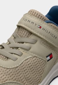 Μπεζ αθλητικό παπούτσι με διχτυωτό επάνω μέρος, επίπεδα κορδόνια και λουράκι με βέλκρο. Διαθέτει λογότυπο Tommy Hilfiger και αντίθετη μπλε επένδυση.