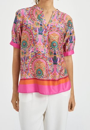 Mujer con una blusa rosa brillante con patrones florales y geométricos multicolores, mangas cortas y pantalones blancos.