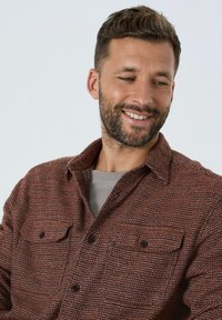 Hubitatud mees, kes naeratab ja vaatab alla, kandes pruuni tekstuuriga nööbiga särk halli T-särgi üle lihtsa tausta ees.