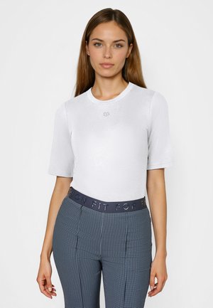 Frau mit langen braunen Haaren, die ein weißes kurzärmeliges Hemd und hoch taillierte marineblaue Hosen mit Muster und einem schwarzen Bund trägt.