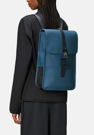 Zaino in pelle blu con chiusura a patta e accenti neri, dotato di spallacci regolabili e un design elegante e minimalista.